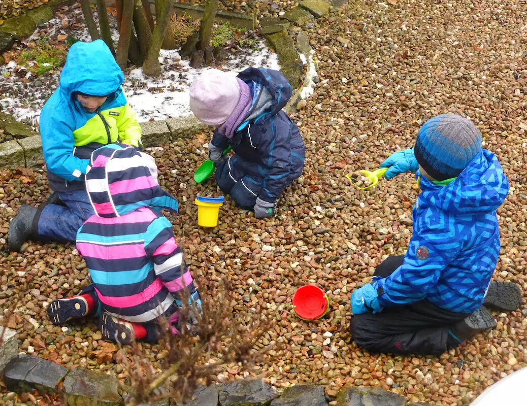 Kleine Kinder in Anoraks spielen im Kies mit Schaufeln und Eimern.