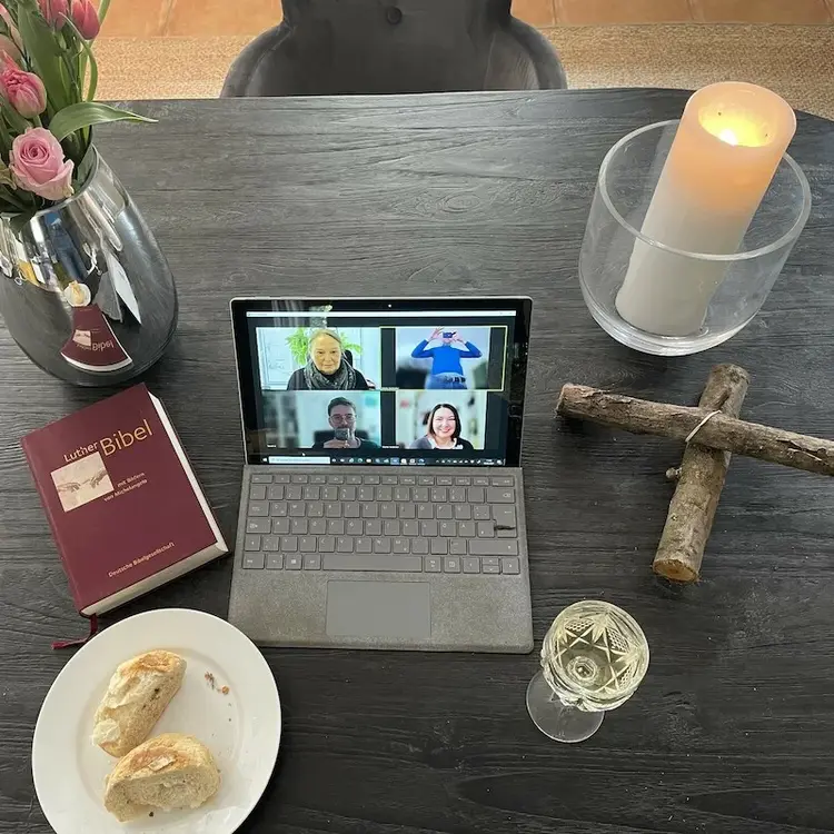 Man sieht einen Tisch mit einem aufgeklappten Notebook und Teilnehmenden einer Zoom Konferenz, daneben einen Teller mit einem Stück Brot, ein gefülltes Weinglas, ein Holzkreuz, eine angezündete Kerze und eine Bibel