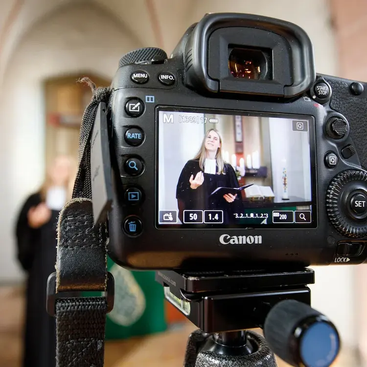 Im Vordergrund ist eine Kamera auf einem Stativ, die eine Pfarrerin in der Kirche filmt